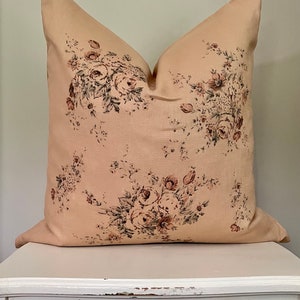 May include: A beige pillow with a floral pattern featuring pink and brown roses and green leaves. The pillow is sitting on a white table.