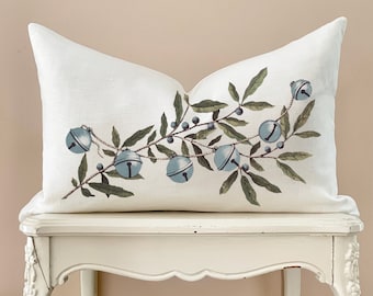 May include: A white linen cushion with a botanical design of blue bells and green leaves. The bells are strung along a vine, creating a festive and elegant aesthetic. This decorative cushion adds a touch of nature-inspired charm to any room.