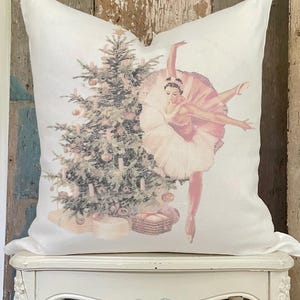 May include: White square pillow featuring a Christmas tree and a ballerina in a pink tutu. The tree is decorated with ornaments and candles. The ballerina is in a graceful pose, with one leg extended. The pillow is on a white table.
