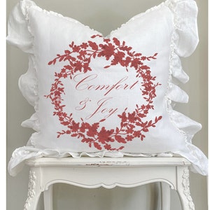 May include: White square pillow with a red floral wreath design and the words "Comfort & Joy." The pillow has ruffled edges. The pillow is sitting on a white table.