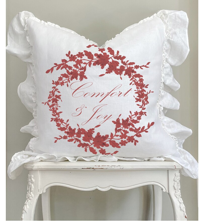 May include: White square pillow with a red floral wreath design and the words "Comfort & Joy." The pillow has ruffled edges. The pillow is sitting on a white table.