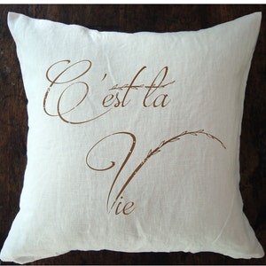 May include: White throw pillow with brown text that reads "C'est la Vie" with a stylized branch design.