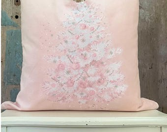 May include: A light pink square pillow featuring a Christmas tree design. The tree is white with pink ornaments and garland. The pillow is resting on a white table with a distressed wood background.