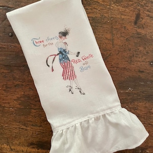 May include: White tea towel with a red, white, and blue illustration of a woman in a patriotic dress. The text "Three cheers for the Red, White and Blue" is printed on the towel.