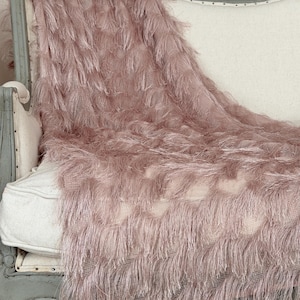 May include: A dusty rose-colored throw blanket with a textured, fringed design draped over a vintage-style chair. The blanket's soft, feathery appearance adds a touch of elegance. The chair has a white cushion and a distressed gray frame.