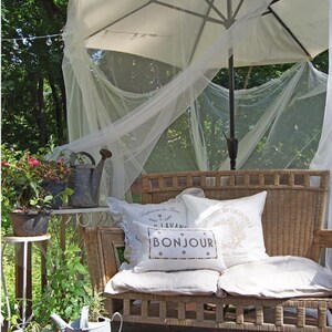May include: A white wicker loveseat with white cushions and pillows. The pillows have French text that reads "Promenade de Fleurs" and "Bonjour". The loveseat is under a white canopy with a white umbrella.