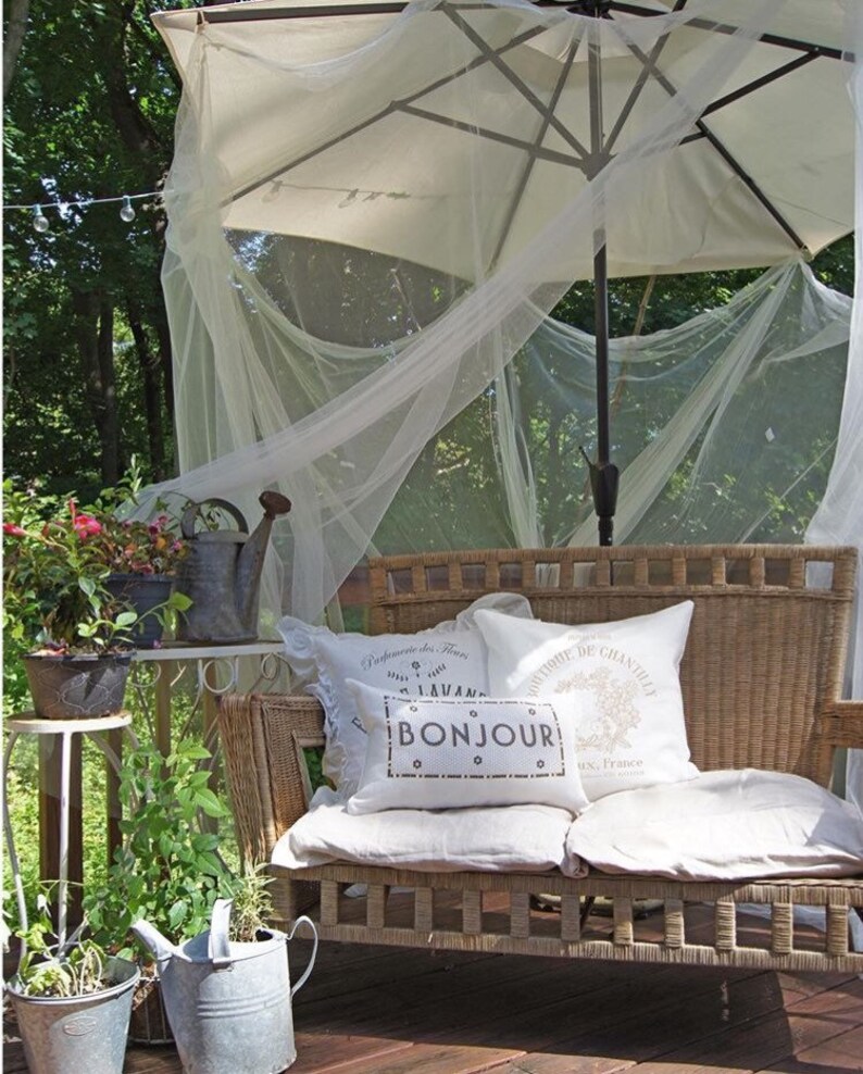 May include: A white wicker loveseat with white cushions and pillows. The pillows have French text that reads "Promenade de Fleurs" and "Bonjour". The loveseat is under a white canopy with a white umbrella.