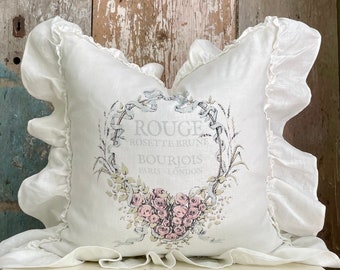 May include: White pillow with a ruffled edge and a printed design featuring a heart of pink roses, a wreath of green leaves, and the text "Rouge Rosette Brune Bourjois Paris London".