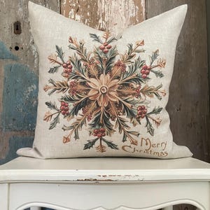 May include: A decorative throw pillow with a beige background. The pillow features a detailed floral design in shades of brown, green, and red, with the words "Merry Christmas" in script. The pillow is placed on a white table.