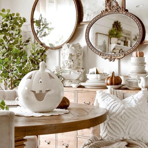Puede incluir: Una calabaza de cer&aacute;mica blanca con una cara sonriente tallada se encuentra sobre una mesa de madera con un mantel blanco. La mesa est&aacute; puesta con un plato blanco y una calabaza marr&oacute;n. El fondo presenta una pared blanca con dos espejos dorados ornamentados y un busto de hombre.