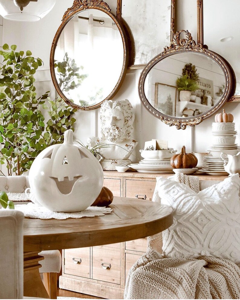 Puede incluir: Una calabaza de cer&aacute;mica blanca con una cara sonriente tallada se encuentra sobre una mesa de madera con un mantel blanco. La mesa est&aacute; puesta con un plato blanco y una calabaza marr&oacute;n. El fondo presenta una pared blanca con dos espejos dorados ornamentados y un busto de hombre.