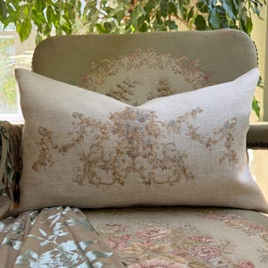 May include: A rectangular lumbar pillow made of light beige linen fabric. The center of the pillow features a printed design of a floral urn surrounded by intricate scrolling foliage and hanging garlands in muted shades of brown, blue, and tan.
