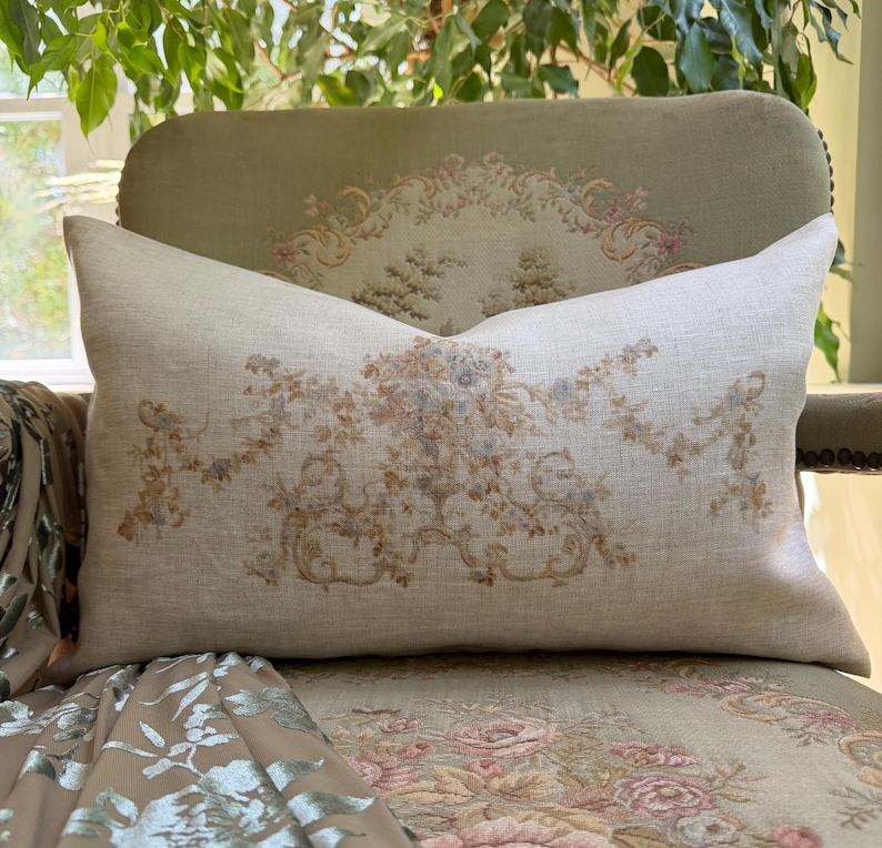 May include: A rectangular lumbar pillow made of light beige linen fabric. The center of the pillow features a printed design of a floral urn surrounded by intricate scrolling foliage and hanging garlands in muted shades of brown, blue, and tan.