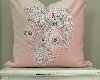 May include: A blush pink square pillow featuring a Christmas ornament design. The design includes silver glittered branches and three pink and white ornaments with red accents. The pillow is resting on a white table.