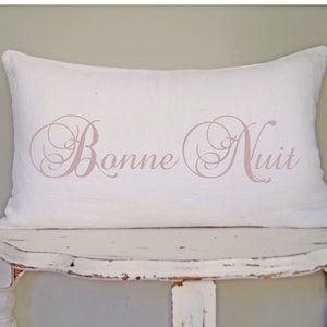 May include: A rectangular white pillow with the words "Bonne Nuit" in a soft pink script. The pillow is made of a textured fabric and rests on a distressed white table, suggesting a bedroom or home decor theme.