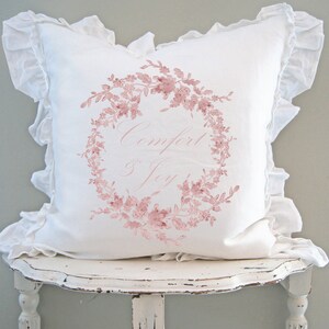 May include: A white decorative pillow with a ruffled edge and a floral wreath design in pink. The words "Comfort & Joy" are written in a delicate script within the wreath. The pillow is placed on a distressed white table.