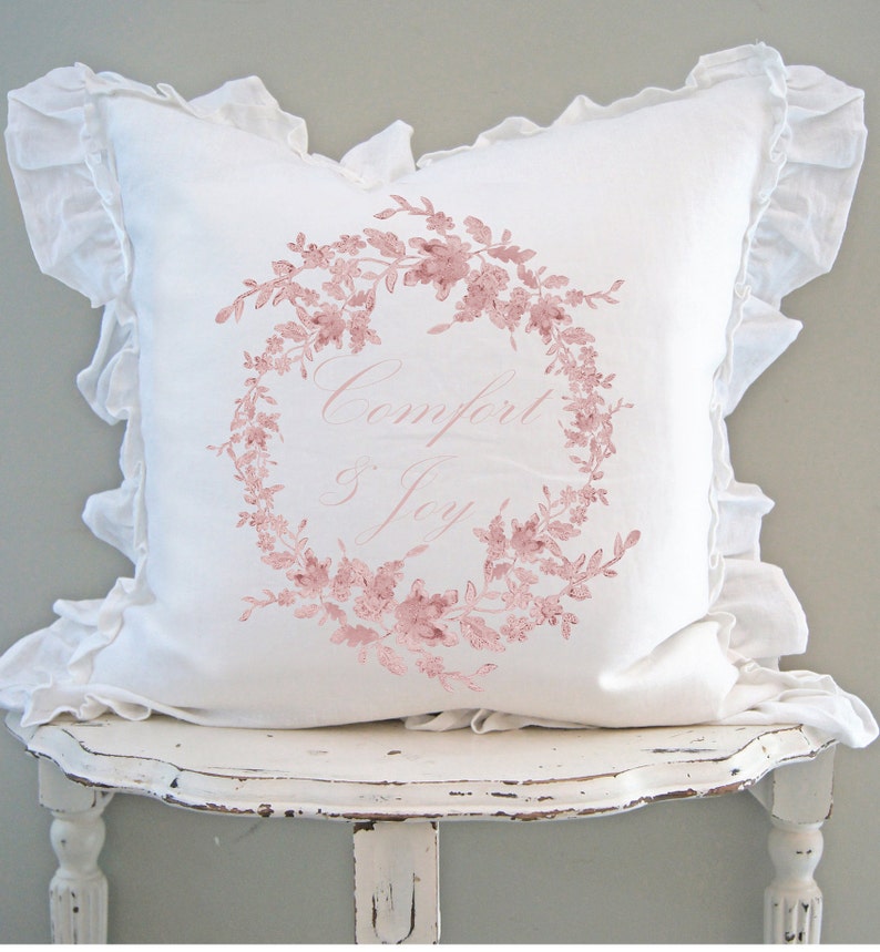 May include: A white decorative pillow with a ruffled edge and a floral wreath design in pink. The words "Comfort & Joy" are written in a delicate script within the wreath. The pillow is placed on a distressed white table.