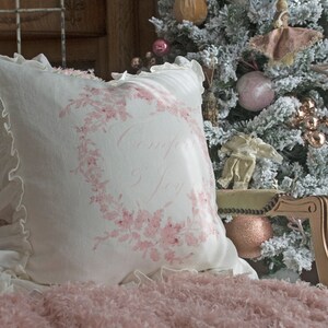 May include: A white pillow with a pink floral wreath design and the words "Comfort Joy" is placed on a pink fluffy blanket. A Christmas tree with ornaments is in the background. The pillow has a ruffled edge.