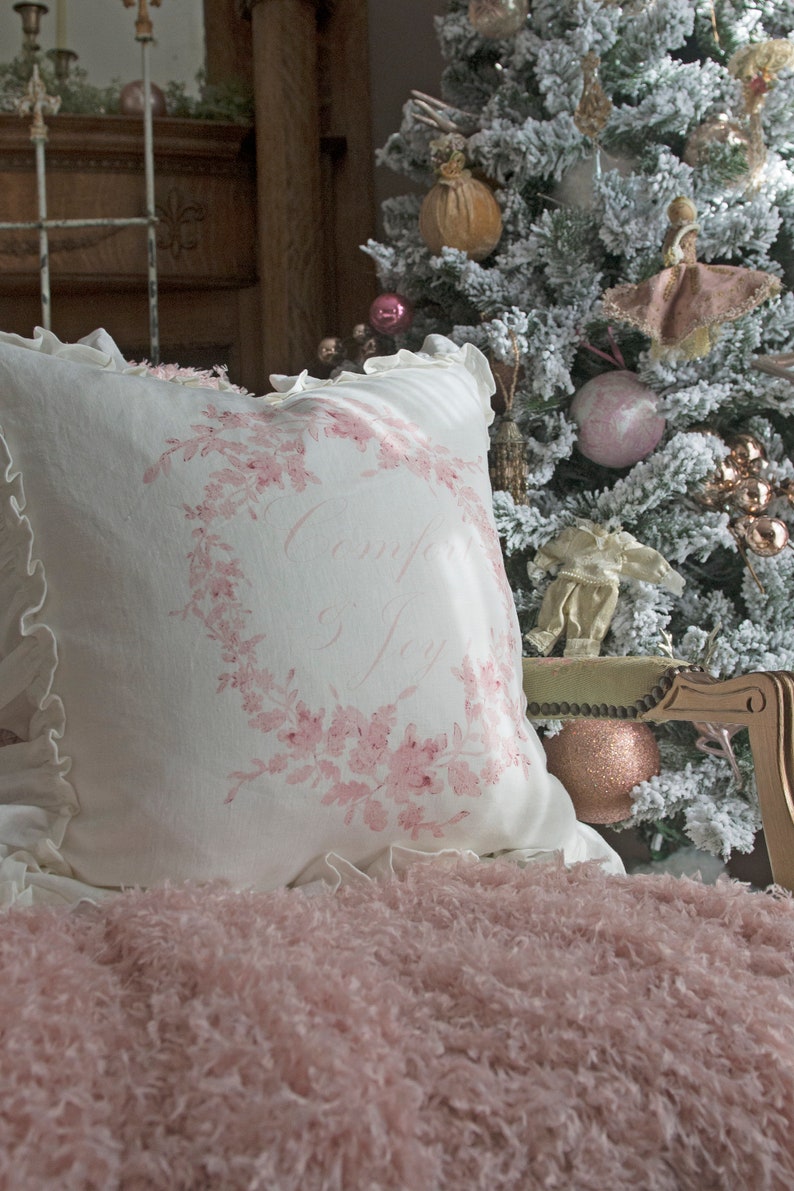 May include: A white pillow with a pink floral wreath design and the words "Comfort Joy" is placed on a pink fluffy blanket. A Christmas tree with ornaments is in the background. The pillow has a ruffled edge.