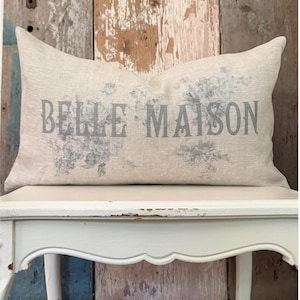 May include: A rectangular beige throw pillow featuring the text BELLE MAISON printed in a grey serif font. The pillow is decorated with a faded grey floral pattern surrounding the lettering, which is centered on the front of the fabric cover.