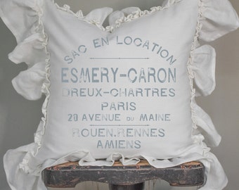 May include: White pillow with ruffled edges and gray text that reads "SAC EN LOCATION ESMERY-CARON DREUX-CHARTRES PARIS 20 AVENUE DU MAINE ROUEN.RENNES AMIENS".
