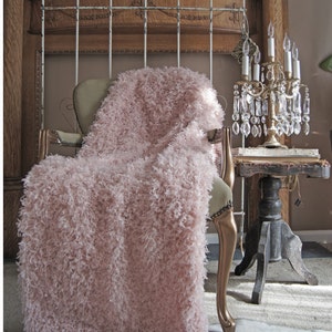 May include: A pink fluffy throw blanket draped over a chair with a gold frame. The chair is in front of a wooden fireplace with a wrought iron grate. A crystal chandelier with candles sits on a small table next to the chair.