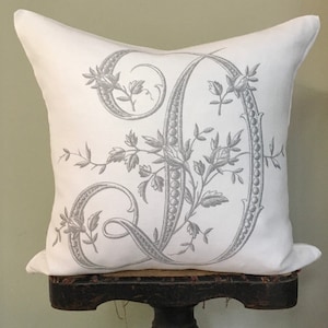 May include: White pillow with a gray embroidered letter 'D' with floral accents.