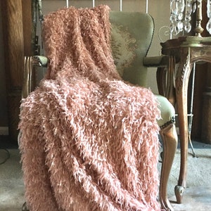 May include: A plush, dusty rose-colored throw blanket draped over a vintage-style armchair. The blanket has a shaggy, textured appearance, adding a touch of luxury. The chair has a light green seat and ornate wooden frame, creating a cozy, inviting aesthetic.