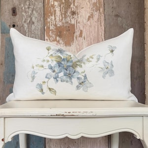 May include: A rectangular throw pillow with an off-white fabric cover featuring a printed botanical illustration of blue flowers, green leaves, and stems centered on the front. The pillow has a smooth texture and a clean, finished edge design.