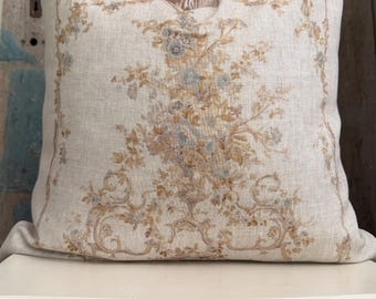May include: A square throw pillow made of light beige fabric featuring a printed design of tan and muted blue floral bouquets arranged in a central cluster, surrounded by a decorative border of scrolling foliage and intricate botanical patterns.