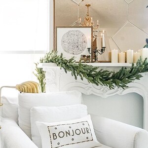 May include: White couch with a white throw pillow that says "BONJOUR" in black lettering. The couch is in front of a white fireplace with a garland of green pine branches.