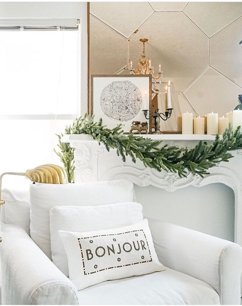 May include: White couch with a white throw pillow that says "BONJOUR" in black lettering. The couch is in front of a white fireplace with a garland of green pine branches.