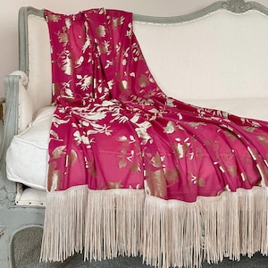 May include: A pink throw blanket with a gold floral pattern and a fringe border. The blanket is draped over a white sofa.