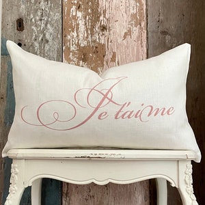 May include: A white linen pillow with a pink cursive "Je t'aime" design. The pillow is sitting on a white stool.