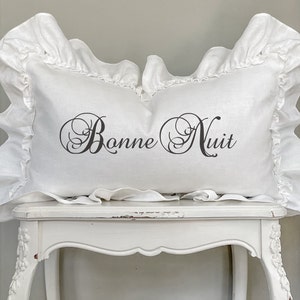 May include: White rectangular pillow with ruffled edges. The pillow features the text "Bonne Nuit" in elegant black script. The pillow is placed on a white, ornate table, creating a soft, inviting aesthetic.