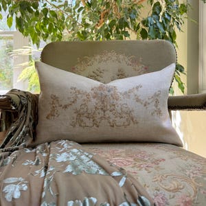 May include: A rectangular throw pillow made of light beige linen fabric featuring a centered, intricate floral and scrollwork pattern in muted shades of brown, tan, and soft blue, with a smooth texture and clean, finished edges.