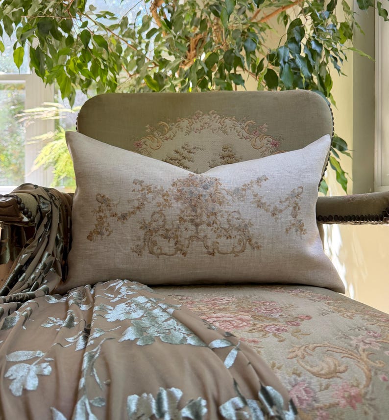 May include: A rectangular throw pillow made of light beige linen fabric featuring a centered, intricate floral and scrollwork pattern in muted shades of brown, tan, and soft blue, with a smooth texture and clean, finished edges.