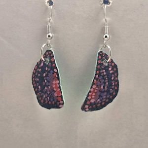 May include: A pair of handmade earrings featuring a butterfly wing design. The earrings have a dark blue background with red and purple dots, and a silver hook. The earrings are a unique accessory.