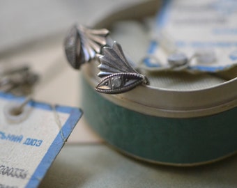 May include: A pair of silver earrings with a fan-shaped design and a small clear stone in the center. The earrings are in a blue and white box with a tag that says "Советская Дио3".