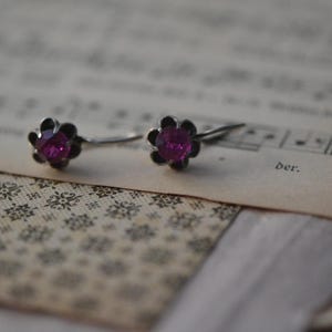 May include: A pair of flower-shaped earrings with a dark silver setting and a bright pink center. The earrings are set on a vintage sheet of music with a floral pattern.