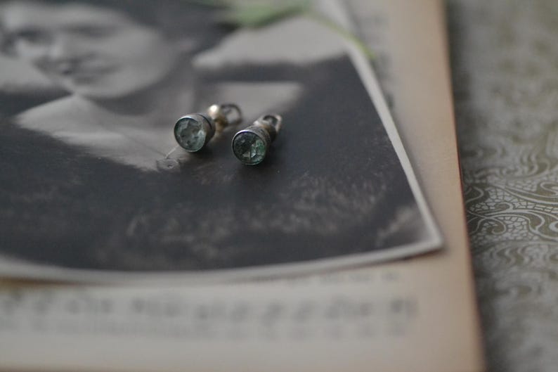 K&ouml;nnte beinhalten: Zwei Vintage-Silberohrringe mit hellblauen Edelsteinen. Die Ohrringe sind vor einer Schwarz-Wei&szlig;-Fotografie einer Frau platziert, die auf einem Vintage-Buch liegt. Die Ohrringe haben ein klassisches, zeitloses Design.