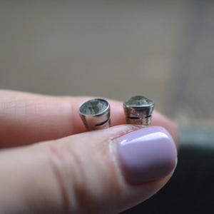 K&ouml;nnte beinhalten: Zwei kleine, silberfarbene Ohrstecker mit einem hellgr&uuml;nen Edelstein. Die Ohrringe werden in einer Hand gehalten. Der Hintergrund ist verschwommen, mit einer neutralen Farbpalette.