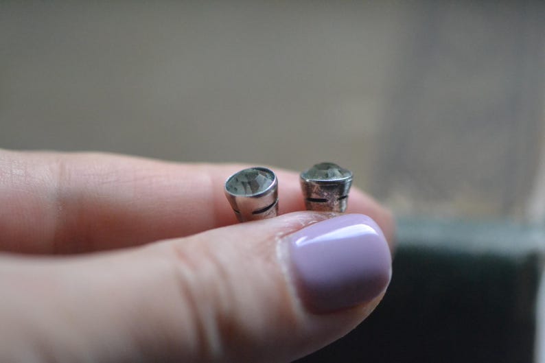 K&ouml;nnte beinhalten: Zwei kleine, silberfarbene Ohrstecker mit einem hellgr&uuml;nen Edelstein. Die Ohrringe werden in einer Hand gehalten. Der Hintergrund ist verschwommen, mit einer neutralen Farbpalette.