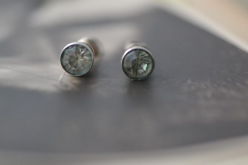 K&ouml;nnte beinhalten: Ein Paar silberfarbene Ohrstecker. Jeder Ohrring hat einen runden, hellblauen Edelstein, der in einer einfachen L&uuml;nette gefasst ist. Die Ohrringe werden vor einem verschwommenen, dunklen Hintergrund gezeigt.