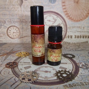 May include: Two amber glass bottles with black roll-on tops and red rubber bands. Both bottles have a brown label with the text "Pain Oil" printed in white. The bottles are sitting on a table with a brown and white patterned surface.
