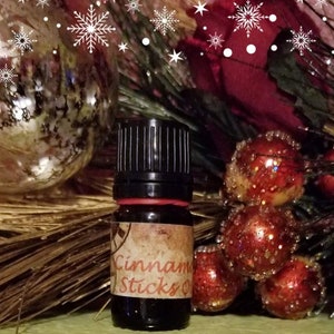 May include: A small, dark brown glass bottle with a black cap and a label that reads "Cinnamon Sticks" in a white font. The bottle is sitting on a brown surface with red and gold ornaments.