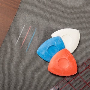 May include: Three triangle-shaped tailor's chalk pieces in white, blue, and orange, with a white, pink, and blue line drawn on a grey fabric surface.