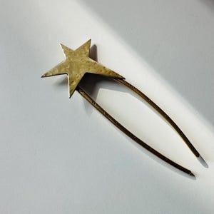 May include: A brass-colored hair pin featuring a five-pointed star design. The star is attached to two long, curved prongs. The hair accessory is designed to secure hair in place and adds a touch of celestial flair.