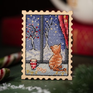Könnte beinhalten: Ein Holz-Kreuzstich-Kunstwerk, das eine Katze zeigt, die aus einem Fenster auf eine verschneite Winterszene blickt. Das Kunstwerk zeigt eine rote Tasse, einen roten Vorhang und einen sternenklaren Nachthimmel. Das Stück hat die Form einer Briefmarke.