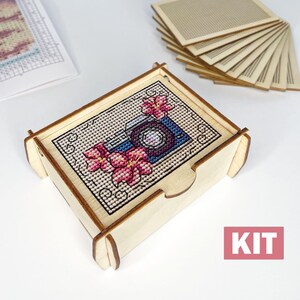 May include: A wooden box with a cross-stitch design of a camera and flowers. The box is labelled "KIT" and is surrounded by a stack of wooden squares.
